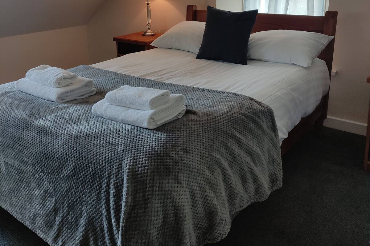 A neatly made bed with white linens and a grey blanket, a bedside lamp, and a window with sheer curtains.