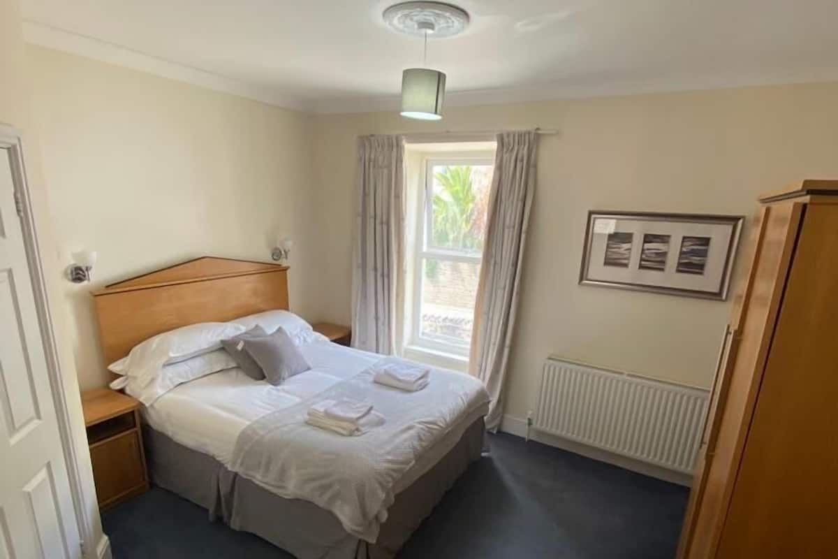 A neatly made bed with white linens, a wooden headboard, and a nightstand with a lamp. There is a window with curtains, a radiator, and a framed picture on the wall.