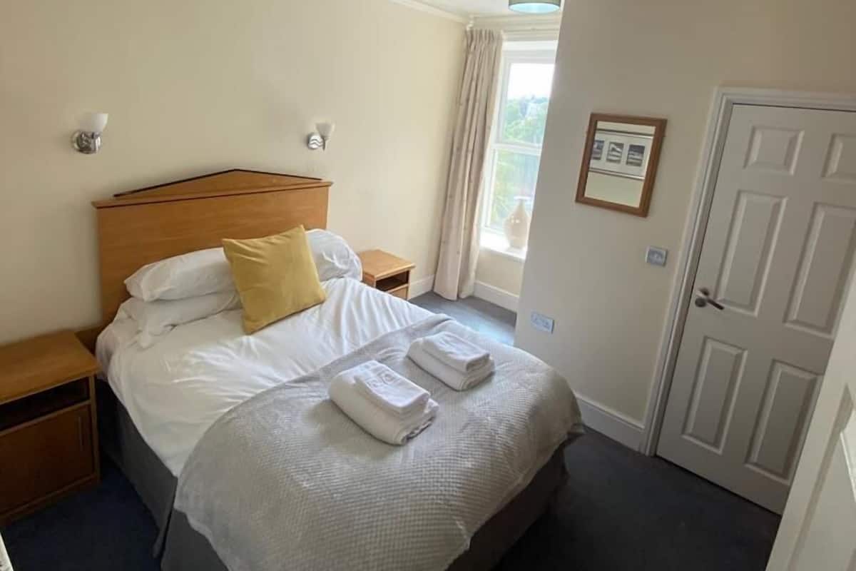 A neatly made bed with white and yellow pillows, a bedside table, a nightstand, a door, and a window with curtains.