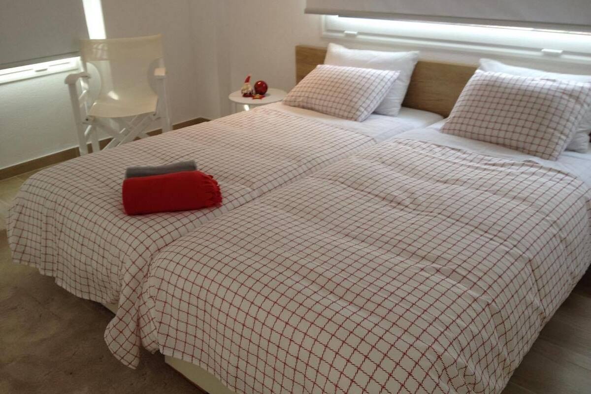 Ein ordentlich bezogenes Bett mit einer karierten Tagesdecke, einem roten Kissen und einem weißen Stuhl in einem Zimmer mit Fenster und Nachttisch, auf dem ein rotes Objekt steht.
