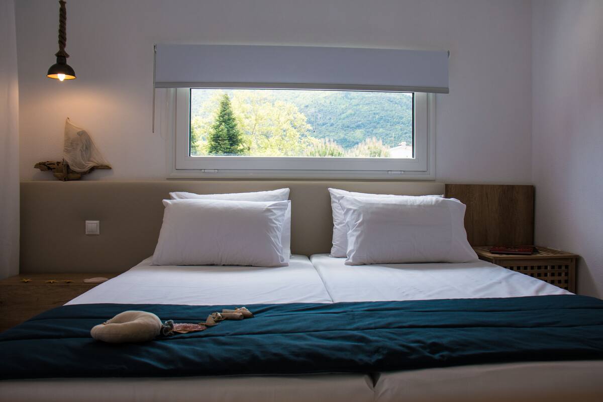 Ein Schlafzimmer mit Bett, Fenster mit Blick auf Bäume, Nachttisch und Lampe.