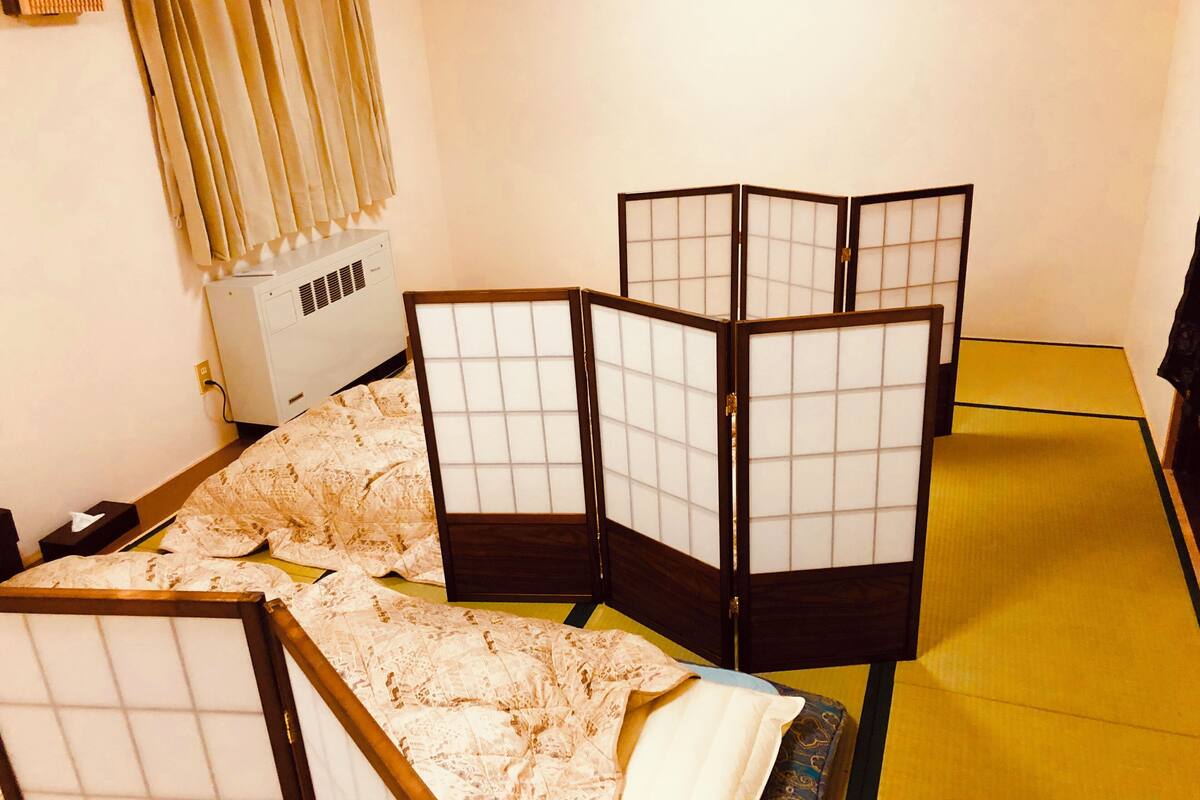 Une pièce avec des tatamis, un lit bas en bois, un paravent japonais traditionnel, un radiateur et une fenêtre avec des rideaux.
