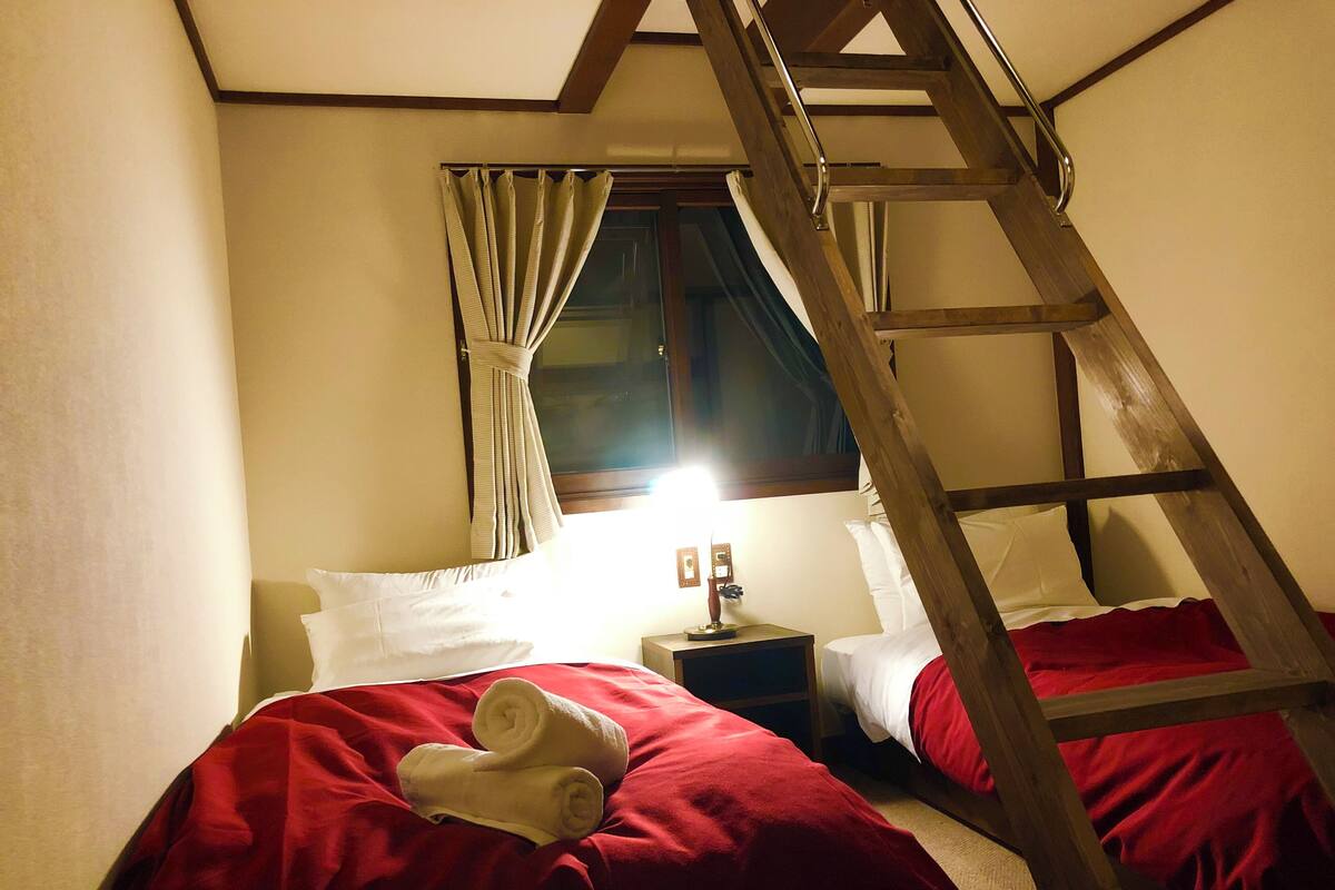 Une pièce avec un lit en mezzanine, une échelle, une table de chevet avec une lampe et une fenêtre avec des rideaux.