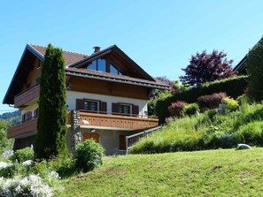 A two-story chalet with a balcony, surrounded by lush greenery and a hillside.