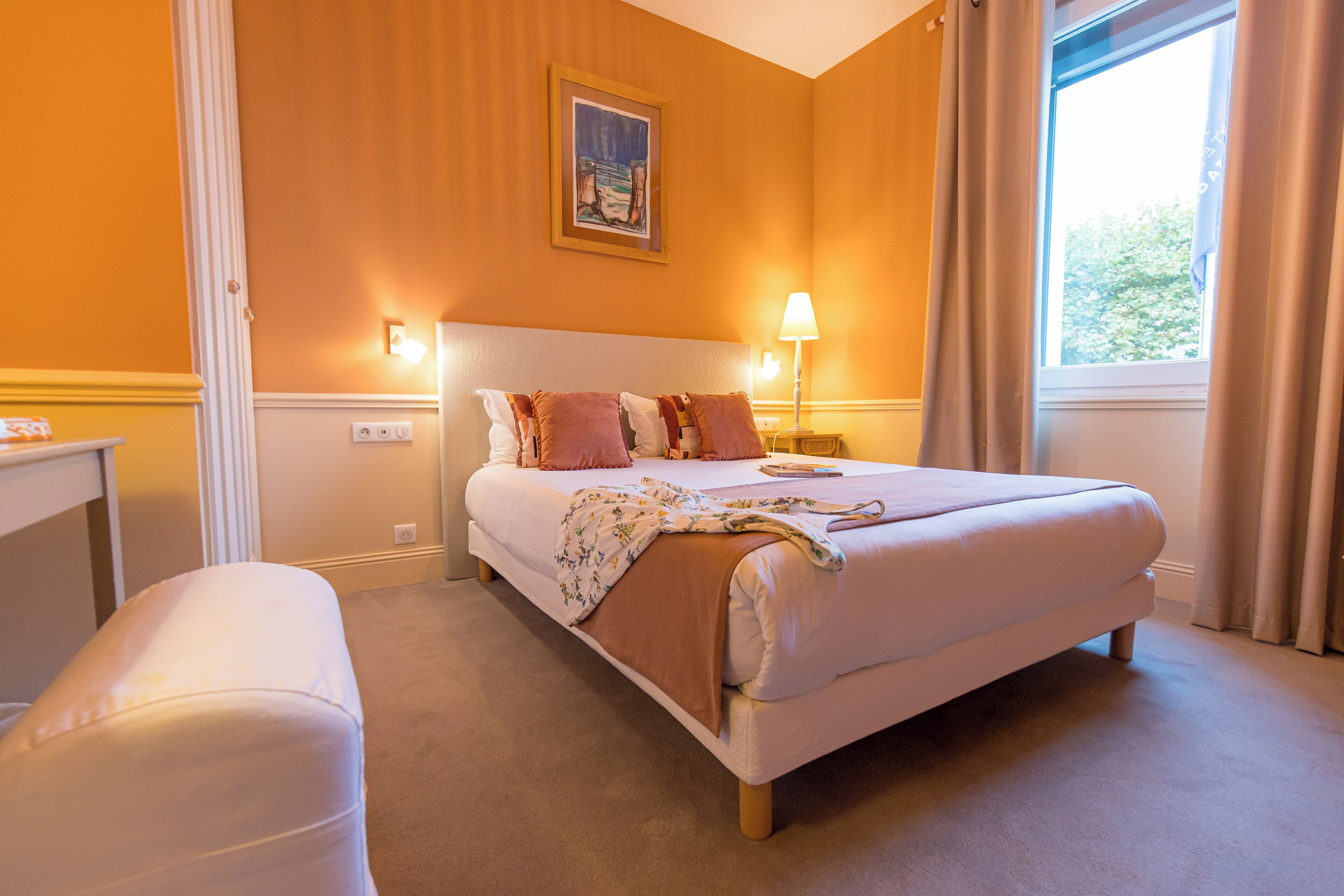 A hotel room with a bed, bedside lamp, window with curtains, and a painting on the wall.