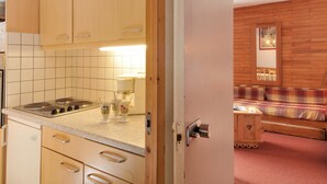 A kitchen with wooden cabinets, a countertop, and a stove. There is a small table with a lamp and a chair. A wooden door leads to a bedroom with a bed and a nightstand. - 2-room apt for 5 in station center with balcony and pool (Plagne Bellecôte)