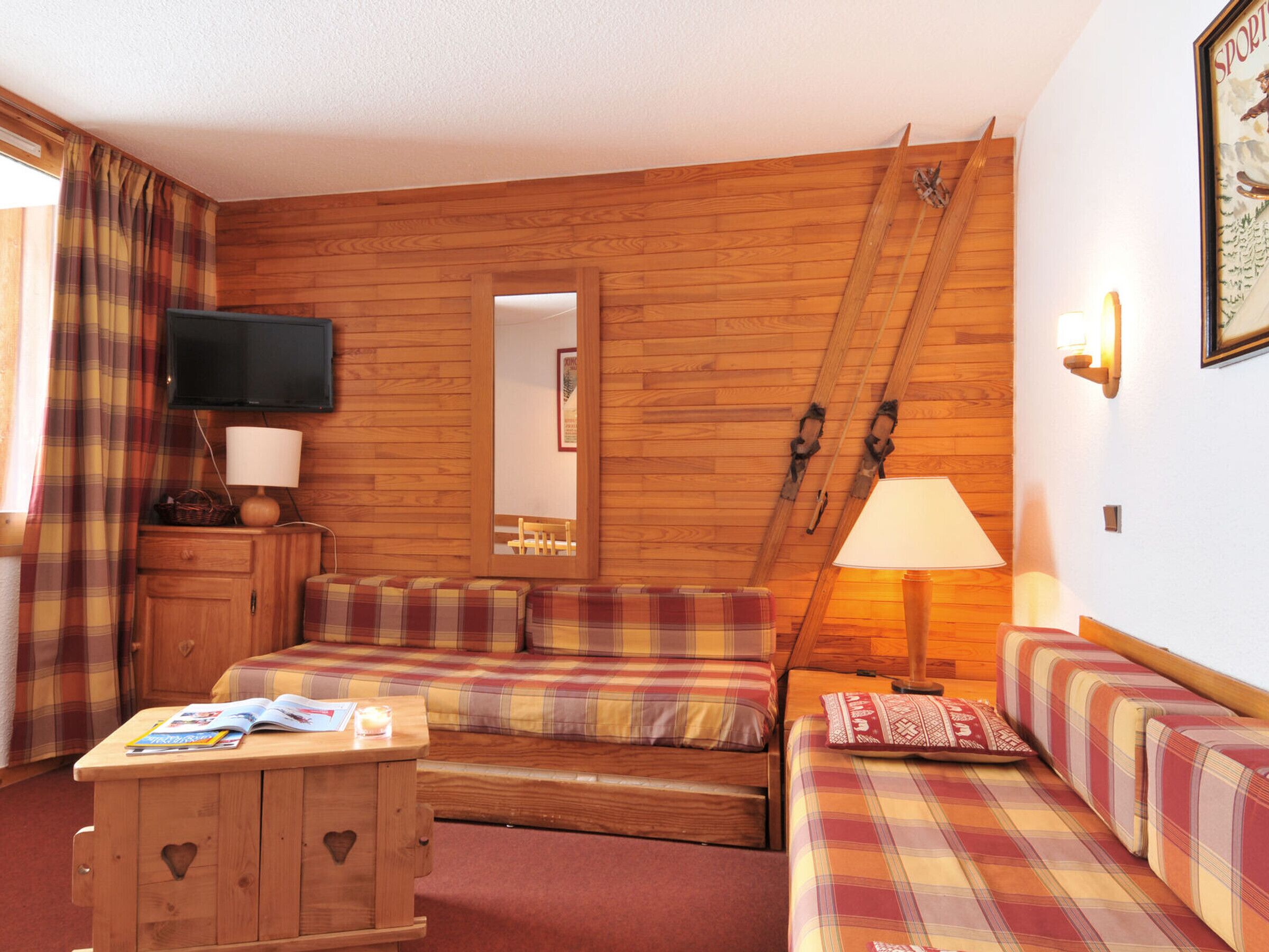A cozy room with wooden paneling, a television, a mirror, skis leaning against the wall, and a sofa with plaid bedding.