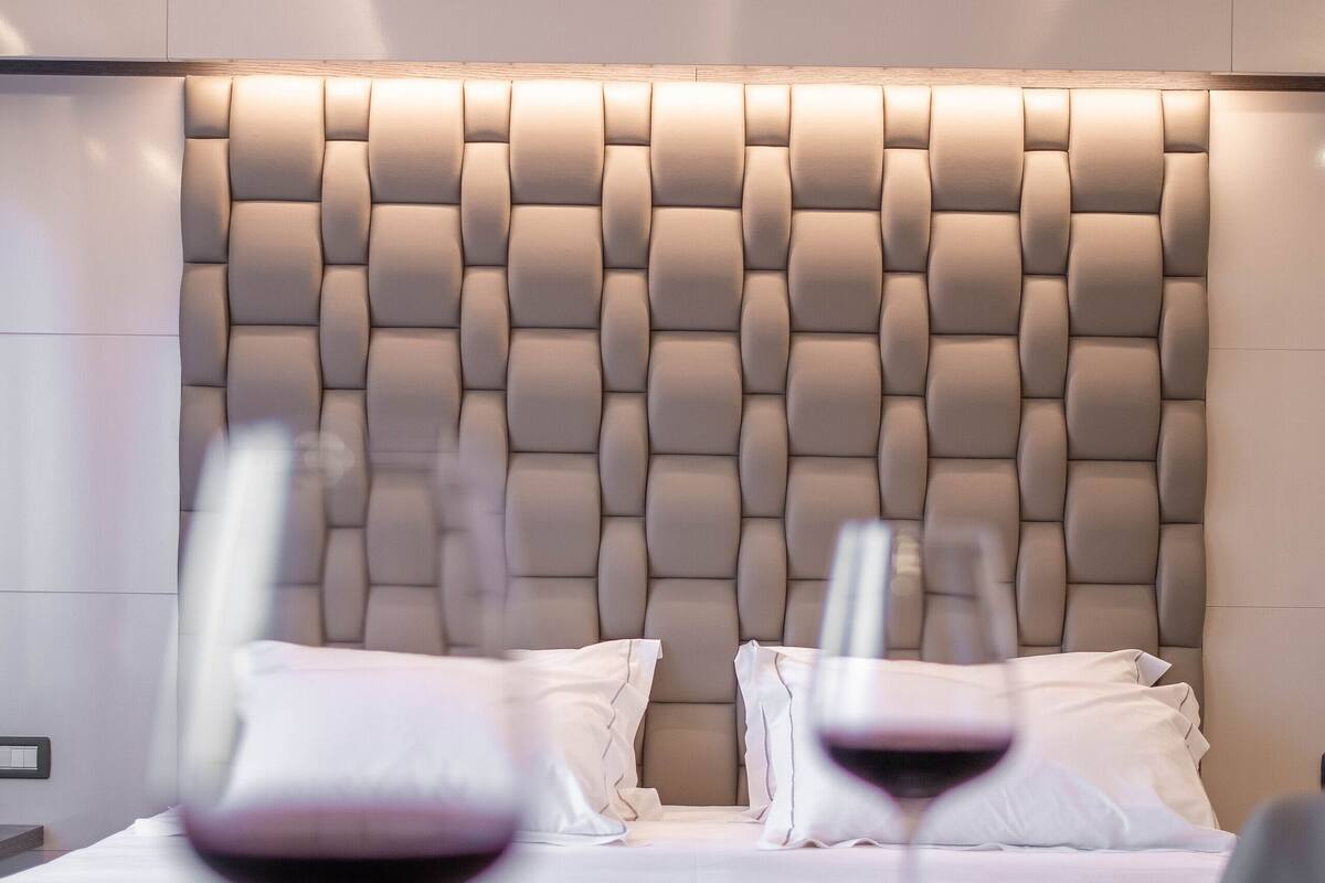 Une chambre d’hôtel moderne avec une tête de lit capitonnée, un lit parfaitement fait et deux verres à vin posés sur une table.