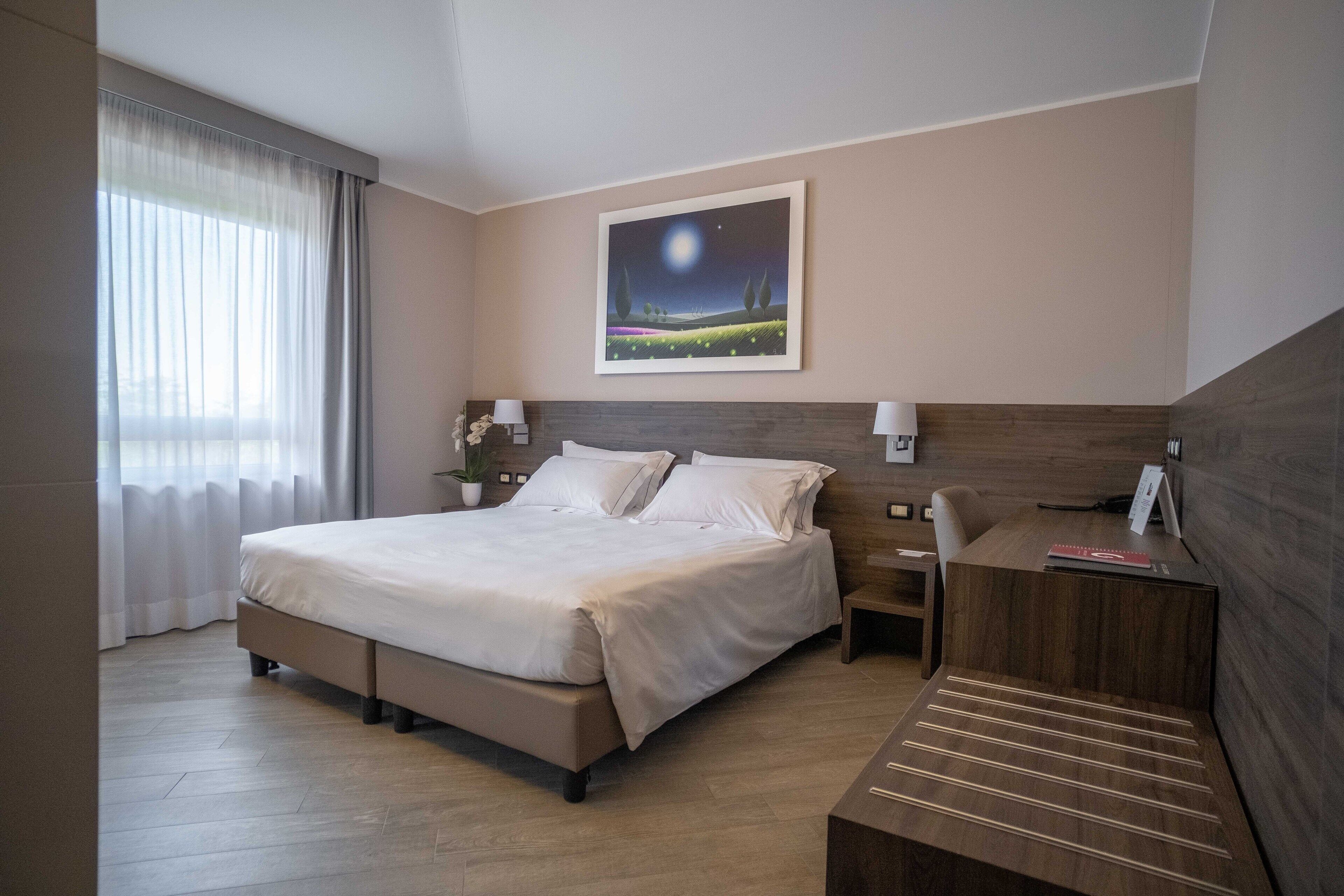A hotel room with a large bed, a desk, a nightstand, and a painting of a night scene on the wall.