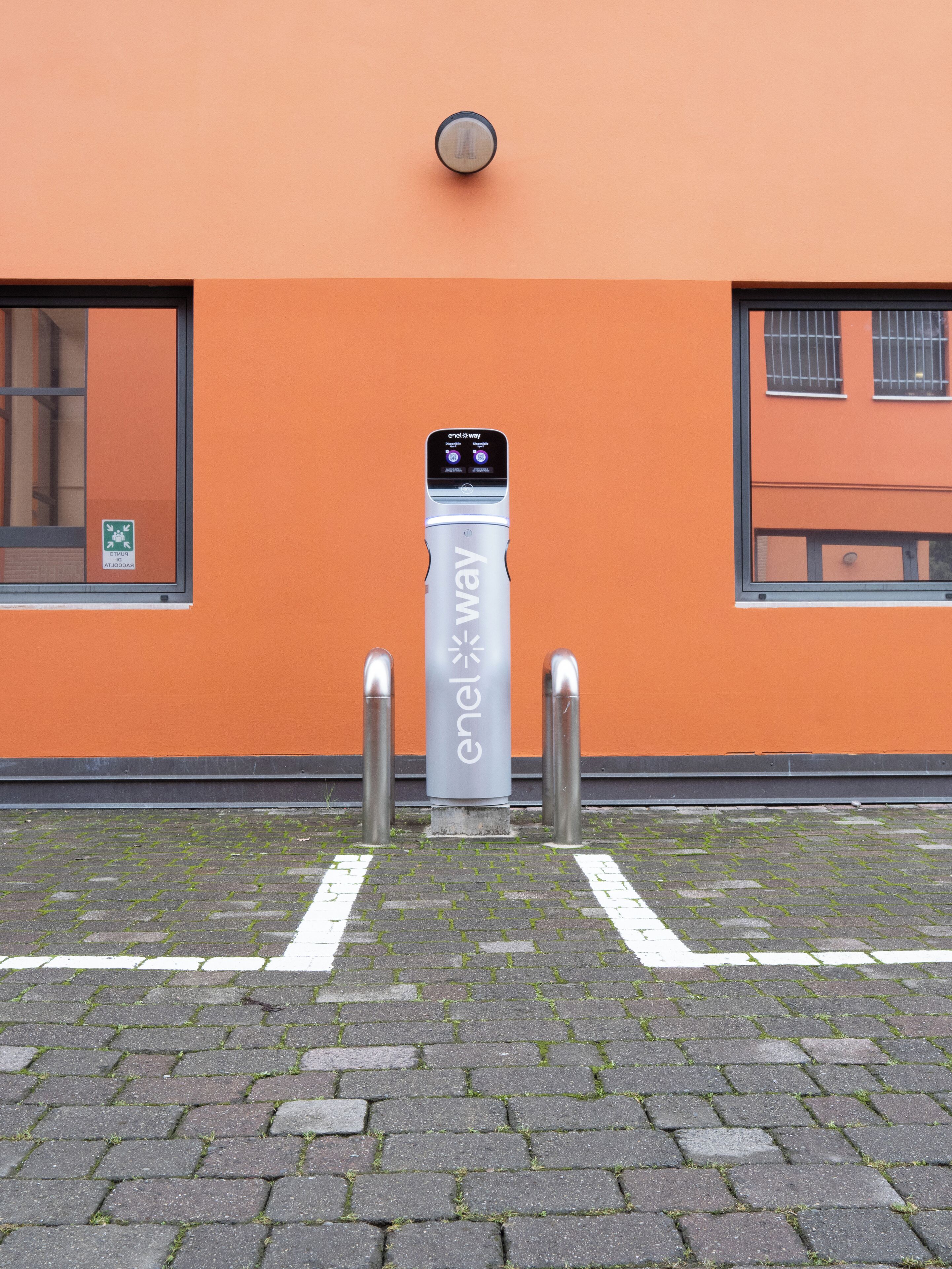 An electric vehicle charging station with a digital display, located in front of an orange building with a window and a door.
