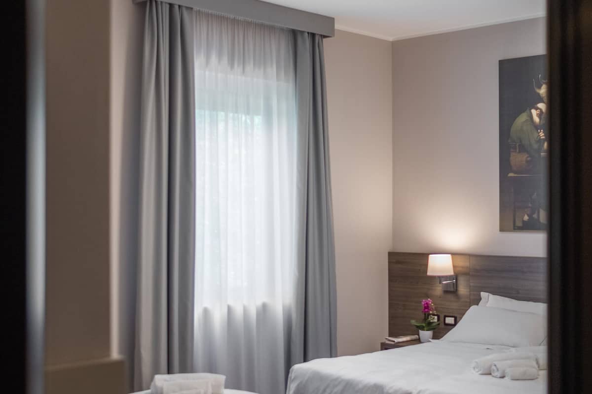 Une chambre d’hôtel avec deux lits, une table de chevet avec une lampe, un tableau au mur et une fenêtre avec des rideaux.