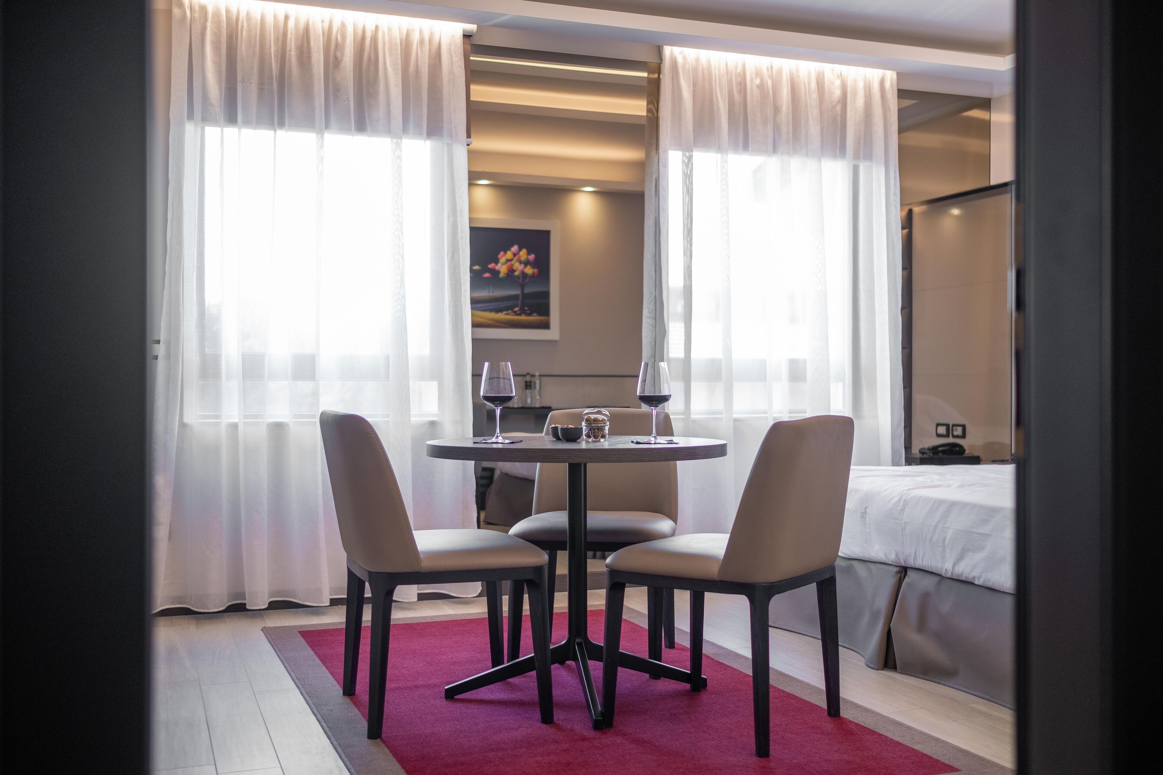 Een hotelkamer met een eethoek, bestaande uit een ronde tafel, vier stoelen en een rood vloerkleed. Er staat een bed met wit beddengoed en een nachtkastje met een lamp. De kamer heeft grote ramen met dunne gordijnen en een schilderij aan de muur.