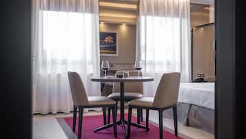 Een hotelkamer met een eethoek, bestaande uit een ronde tafel, vier stoelen en een rood vloerkleed. Er staat een bed met wit beddengoed en een nachtkastje met een lamp. De kamer heeft grote ramen met dunne gordijnen en een schilderij aan de muur.