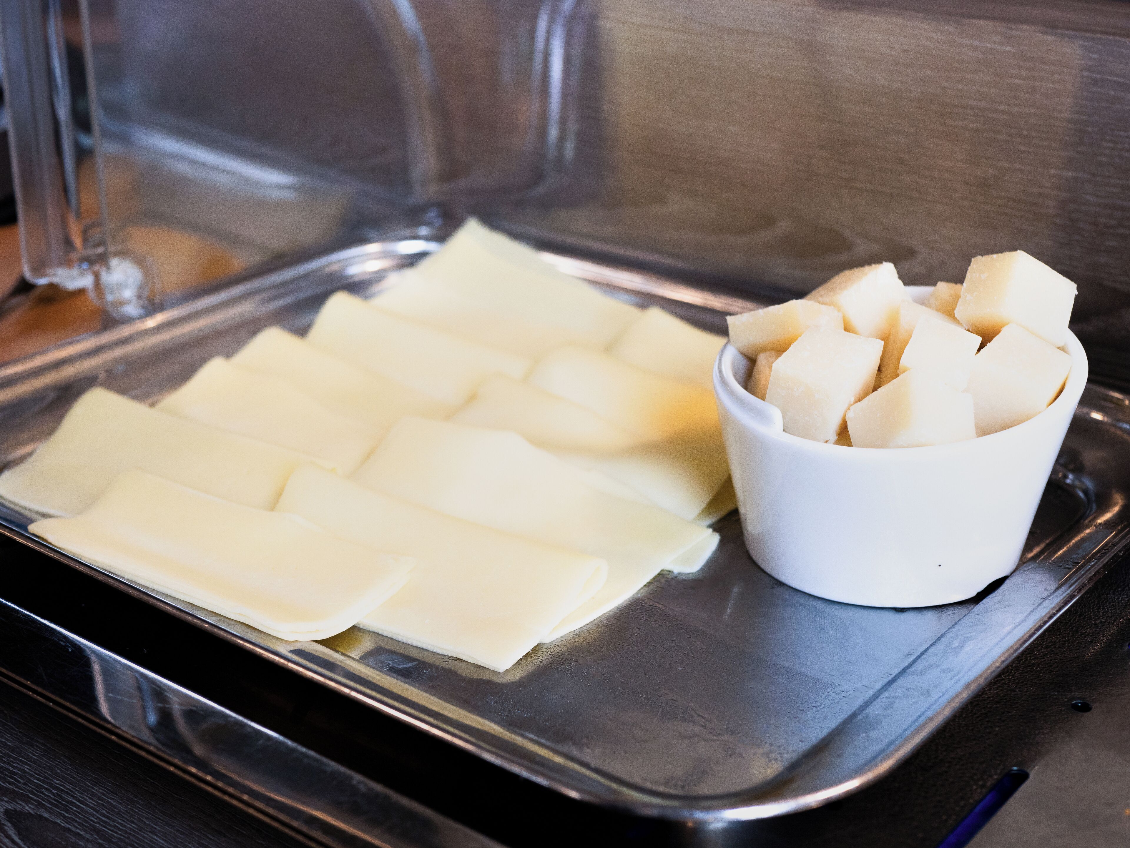 Una bandeja de acero inoxidable con alimentos rectangulares ordenados y un recipiente blanco pequeño con cubos de comida.