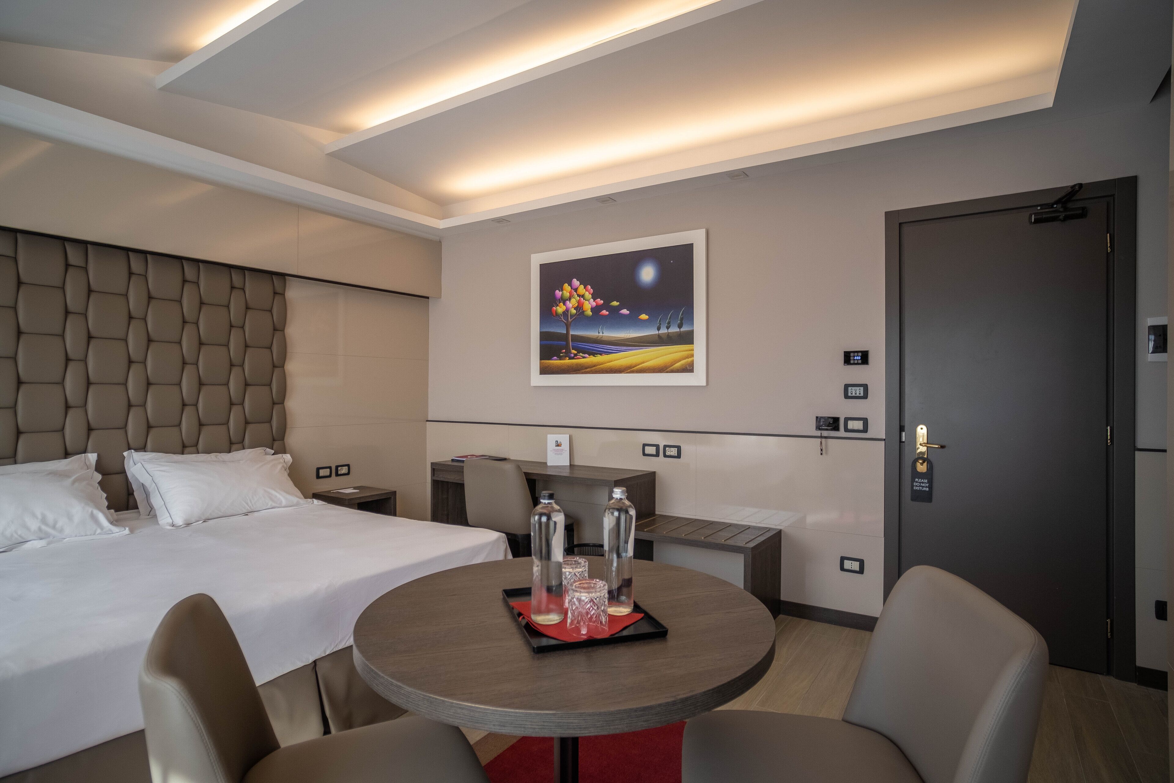 Ein Hotelzimmer mit einem Bett, einem runden Tisch mit einem Tablett mit Gläsern und Flaschen, einem Bild an der Wand und einer Tür mit Schloss.