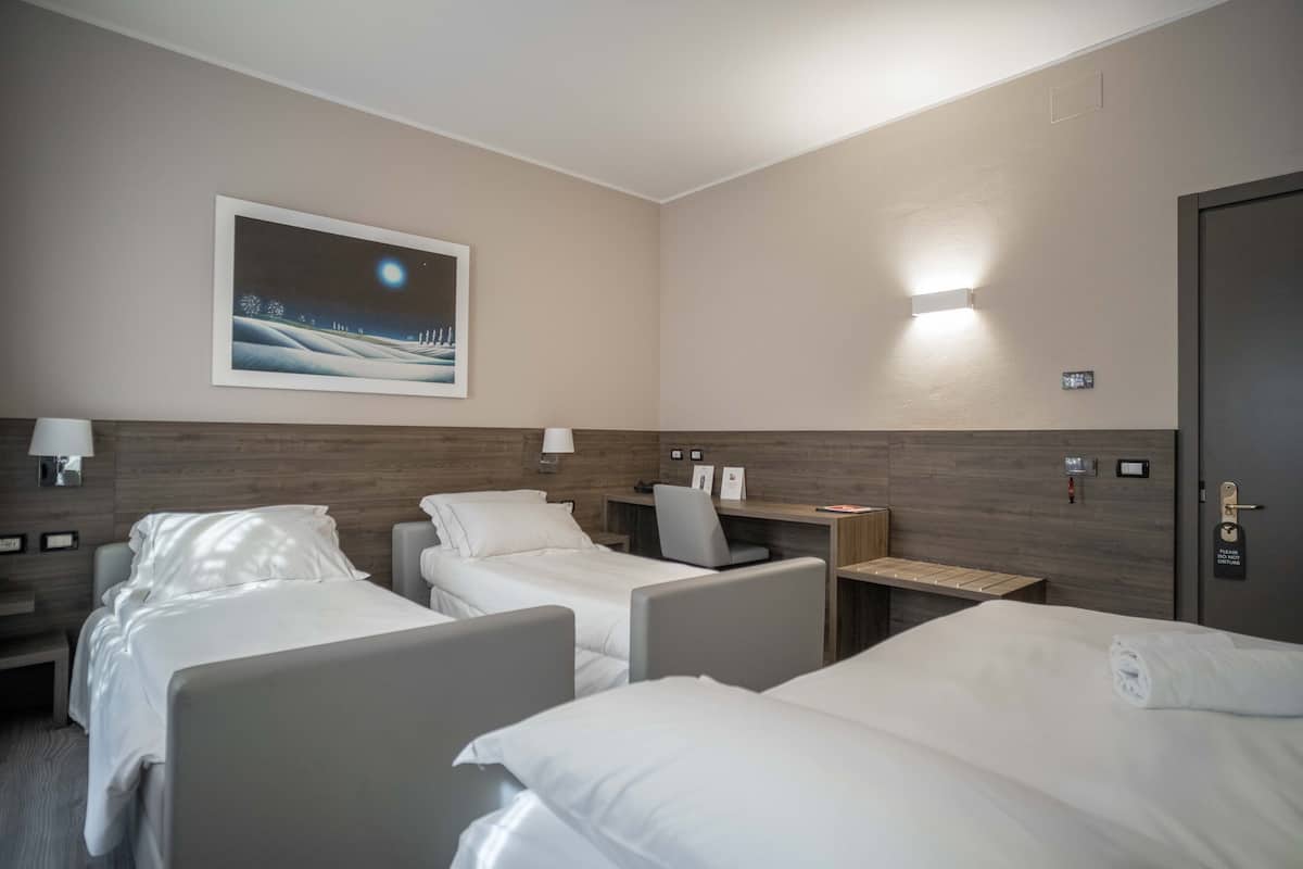 Une chambre d’hôtel avec deux lits, un bureau, une chaise, une table de chevet et une lampe fixée au mur.