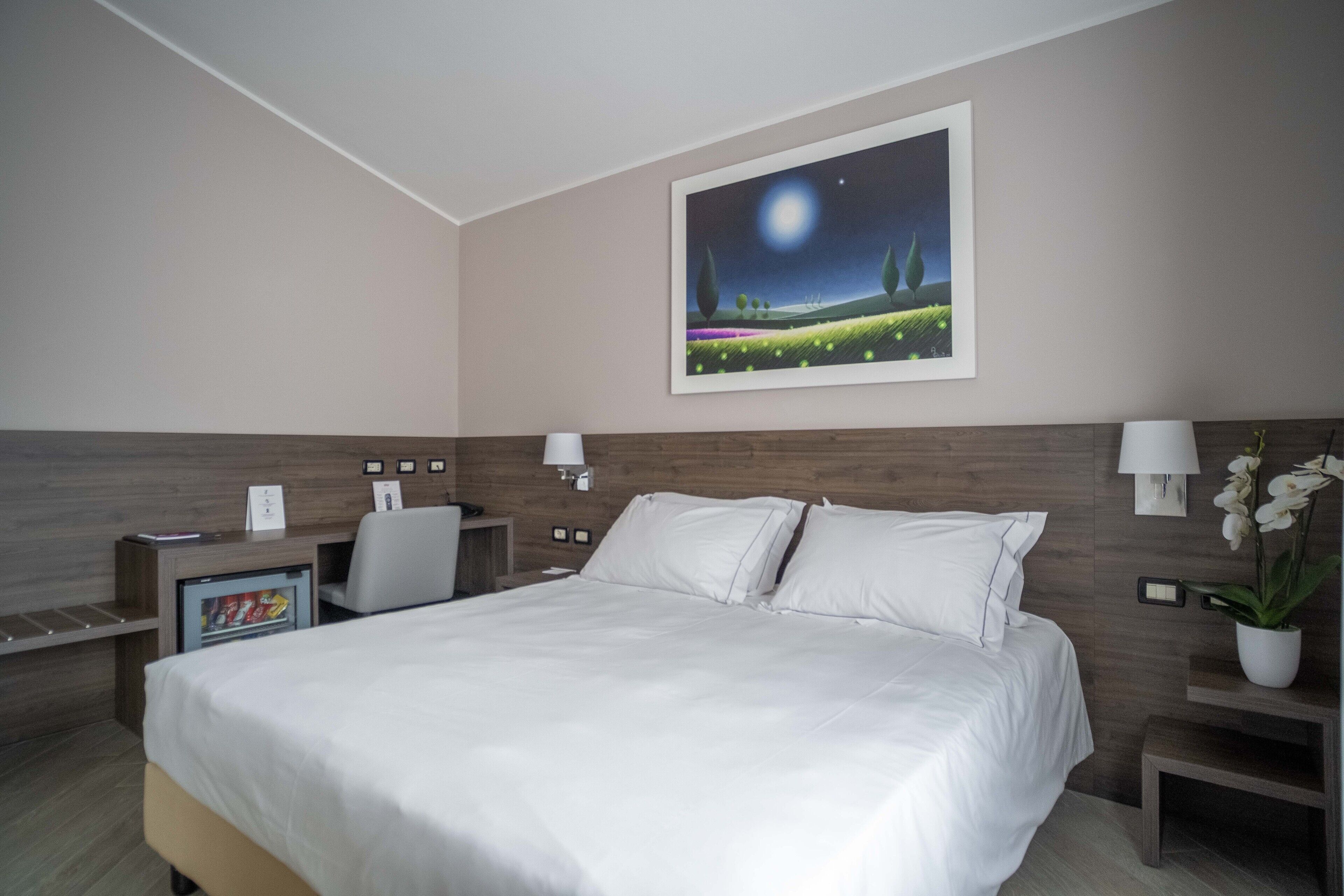 A hotel room with a large bed, a desk with a computer, a chair, a nightstand with a lamp, and a framed picture on the wall.