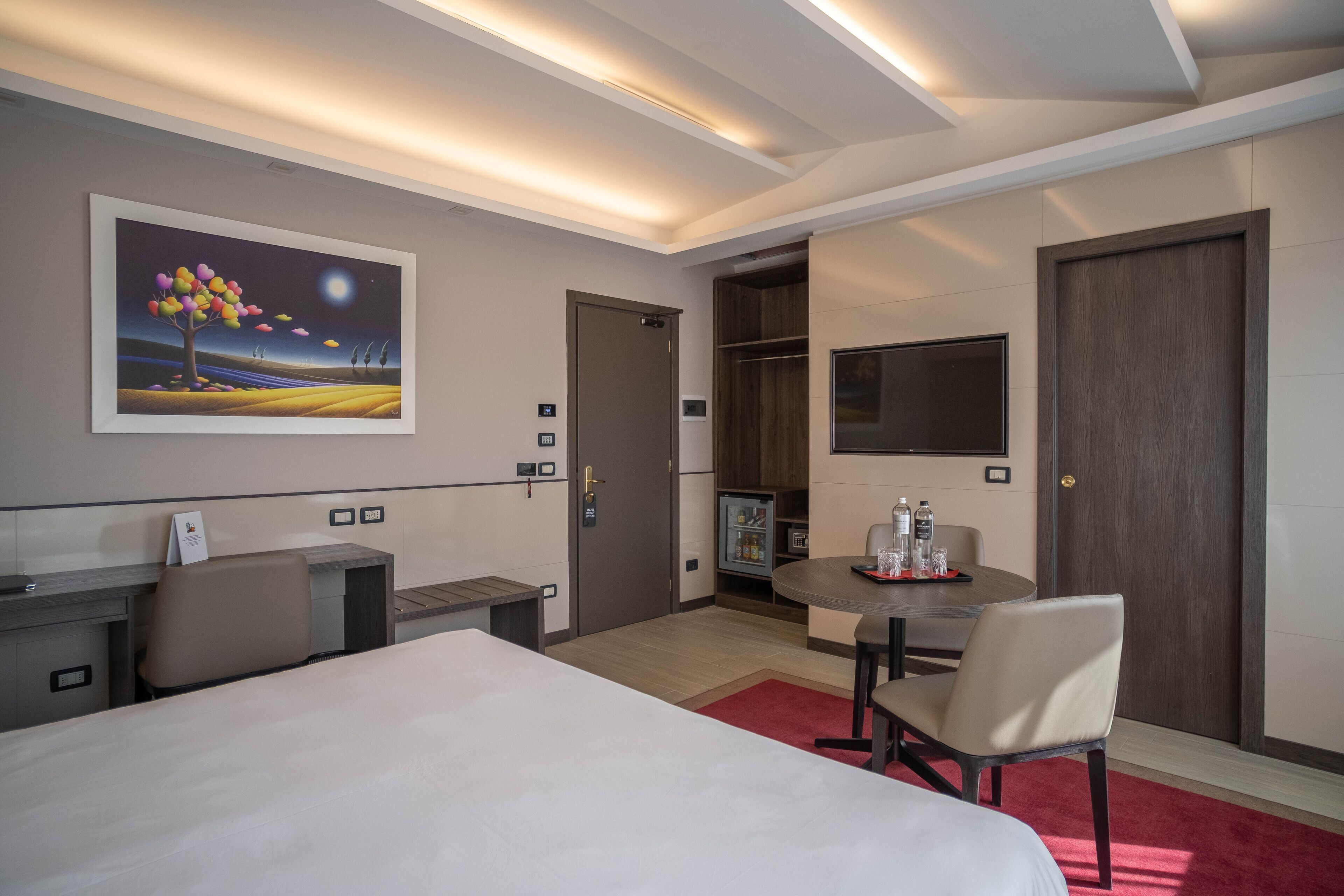 A hotel room with a bed, desk, chair, and a dining table with chairs. There is a painting on the wall, a TV mounted on the wall, and a door leading to another room.