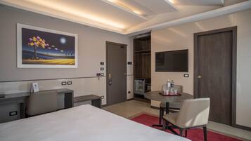 A hotel room with a bed, desk, chair, and a dining table with chairs. There is a painting on the wall, a TV mounted on the wall, and a door leading to another room.
