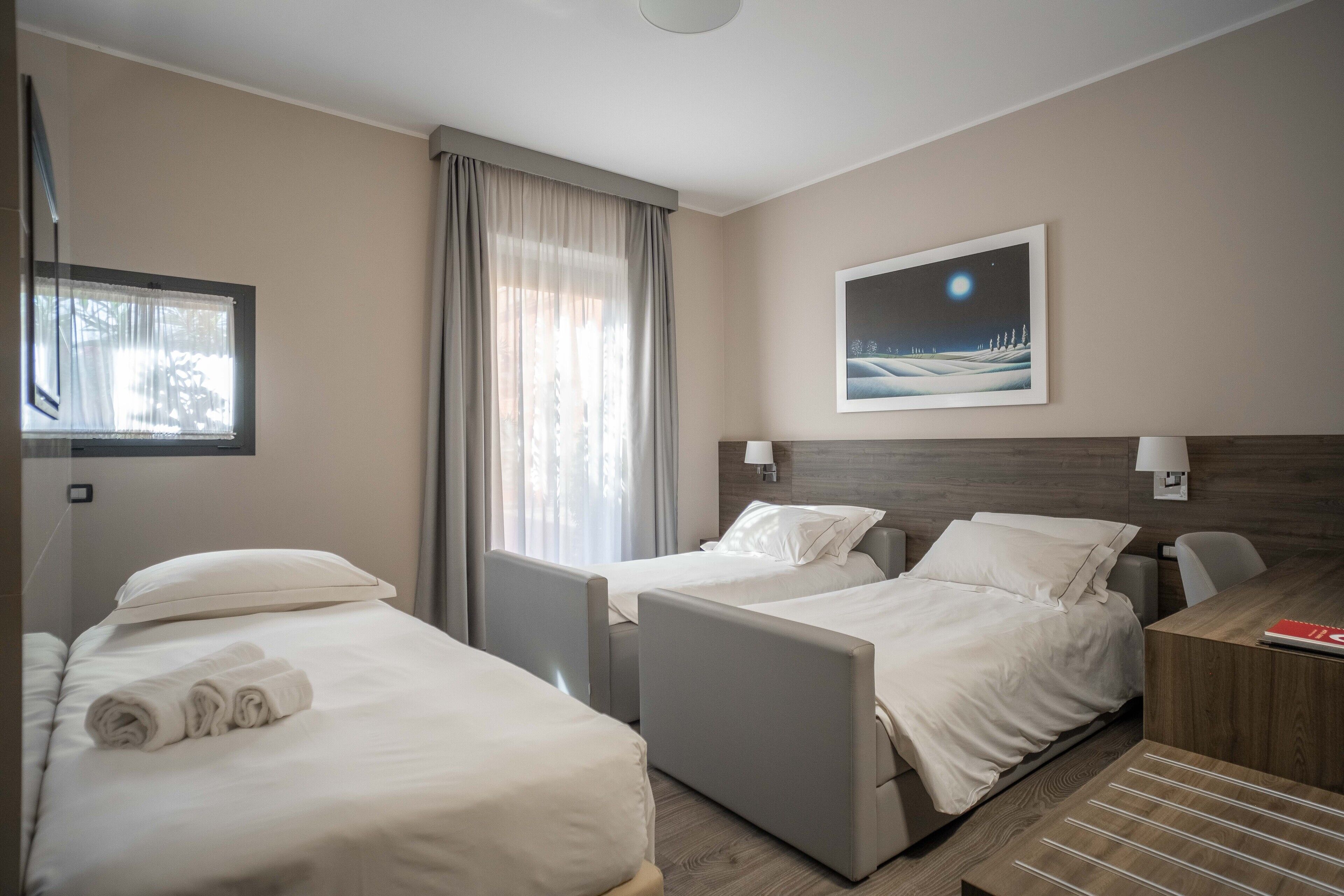 Een hotelkamer met twee bedden, een bureau en een nachtkastje. Er is een raam met gordijnen, een schilderij aan de muur en een lamp op het nachtkastje.