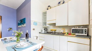 Een kleine keuken met witte kastjes, een gasfornuis, een magnetron en een spoelbak. Er is een eethoek met een tafel voor twee personen, een vaas met bloemen en een fles wijn.