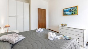 Een net opgemaakt bed met een grijze deken, twee opgerolde handdoeken en een tijdschrift. Er is een witte dressoir met een schilderij erboven, een houten deur en een witte garderobe met een lamp op een bijzettafeltje.