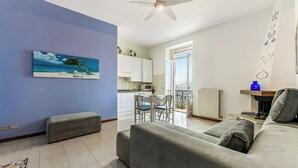 Een woonkamer met een grijze bank, een houten bankje, een plafondventilator en een schilderij van een zandstrand.