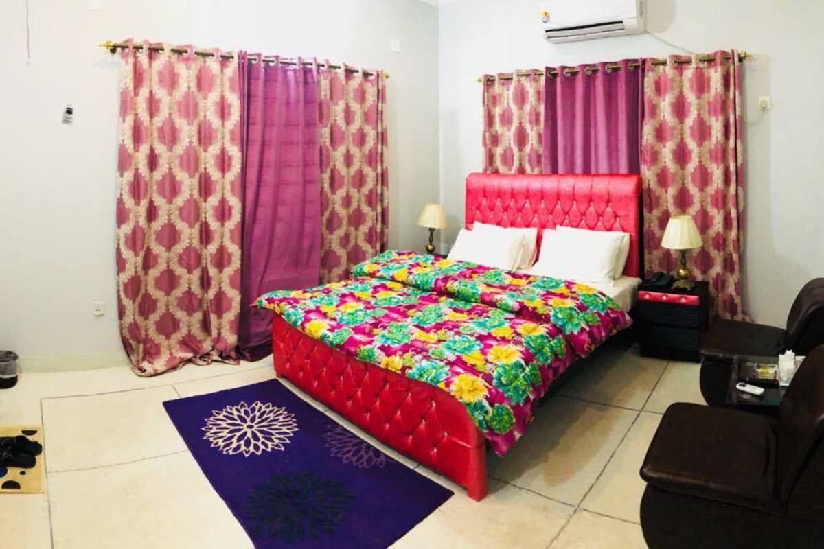A bedroom with a red bed, patterned curtains, a TV, a desk, and a chair.