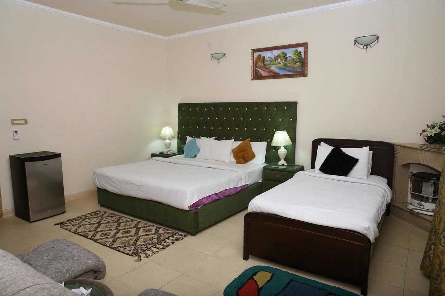 A hotel room with two beds, a green headboard, a small heater, a nightstand with a lamp, a painting on the wall, and a small speaker.