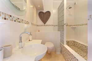 A bathroom with a large white sink, a toilet, a shower with mosaic tiles, and a heart-shaped decoration on the wall. - Neptune Isle 214 (Cape Town)