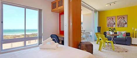 A hotel room with a bed, a view of the beach, a dining area with yellow chairs, and a sofa.