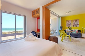 Une chambre d’hôtel avec un lit, une vue sur la plage, un coin repas avec des chaises jaunes et un canapé.