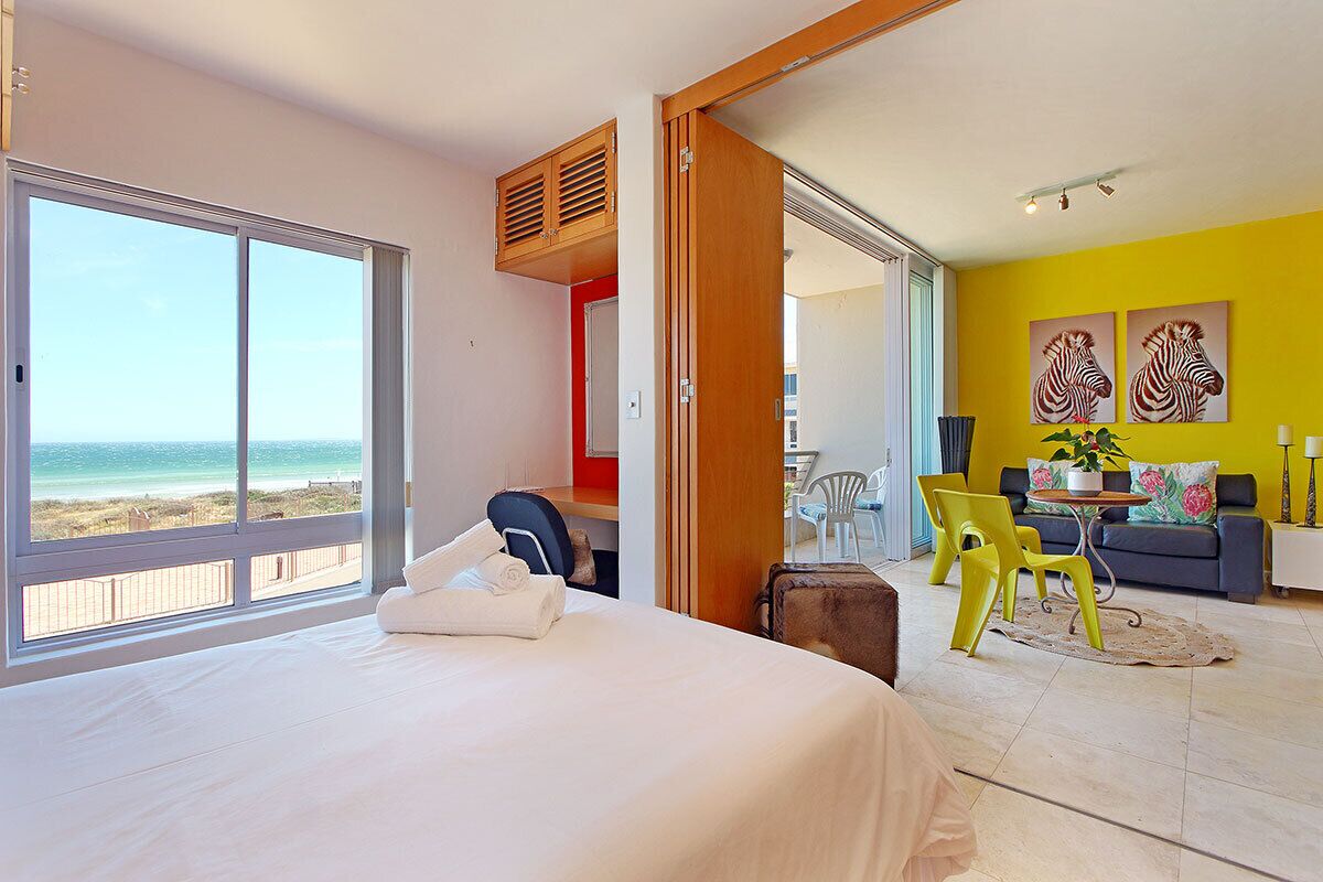 Une chambre d’hôtel avec un lit, une vue sur la plage, un coin repas avec des chaises jaunes et un canapé.