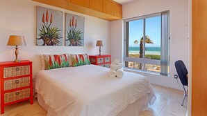 A bedroom with a bed, two nightstands, a red side table, a window with a view of the beach, and artwork on the wall. - Neptune Isle 214 (Cape Town)