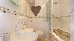 A bathroom with a large white sink, a toilet, a shower with mosaic tiles, and a heart-shaped decoration on the wall. - Neptune Isle 214 (Cape Town)