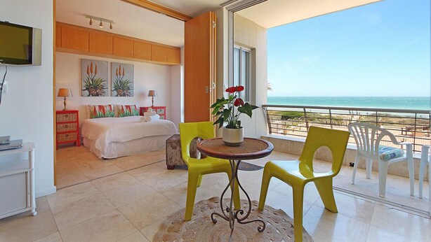 A hotel room with a bed, a small table with chairs, a TV, and a balcony with a view of the sea. - Neptune Isle 214 (Cape Town)