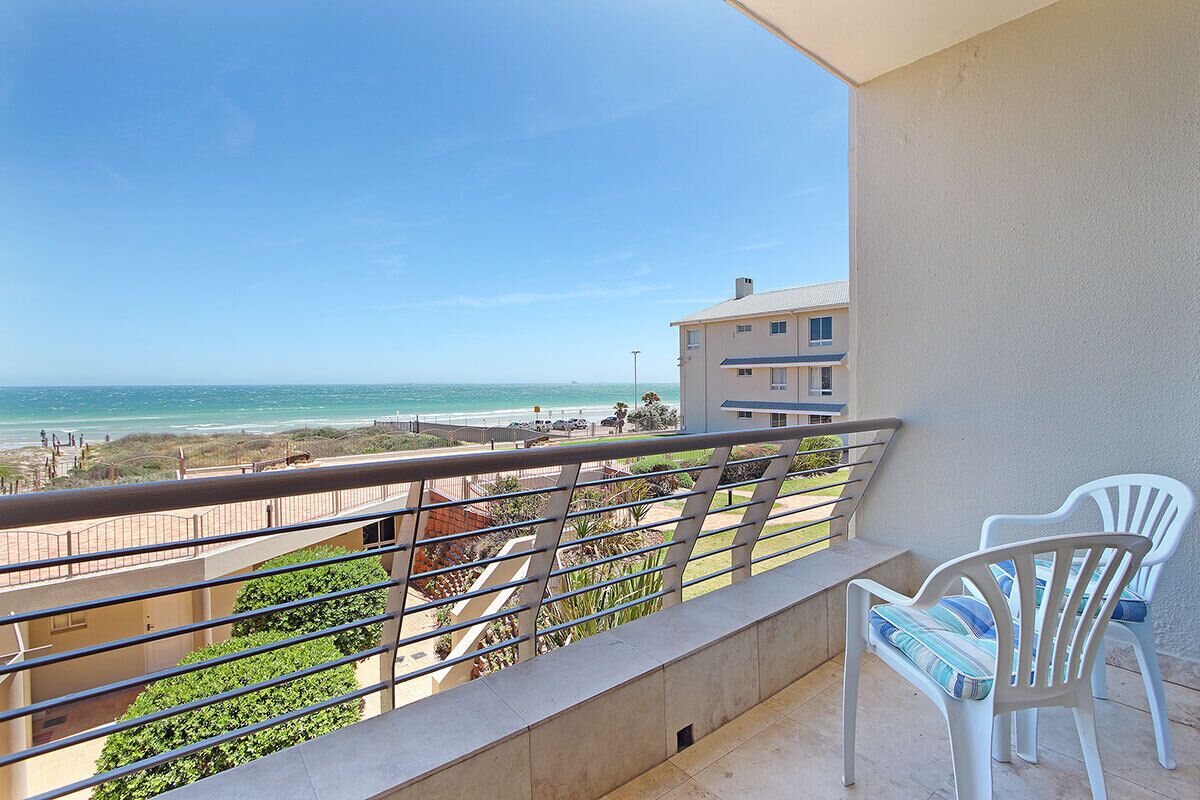 A balcony with a view of the beach, white chairs, and a railing.
