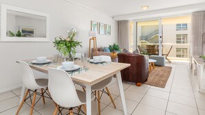A modern living room with a dining area, featuring a wooden dining table, white chairs, a brown sofa, and a large window with a view of the outside. - Lagoon Beach 231 (Cape Town)