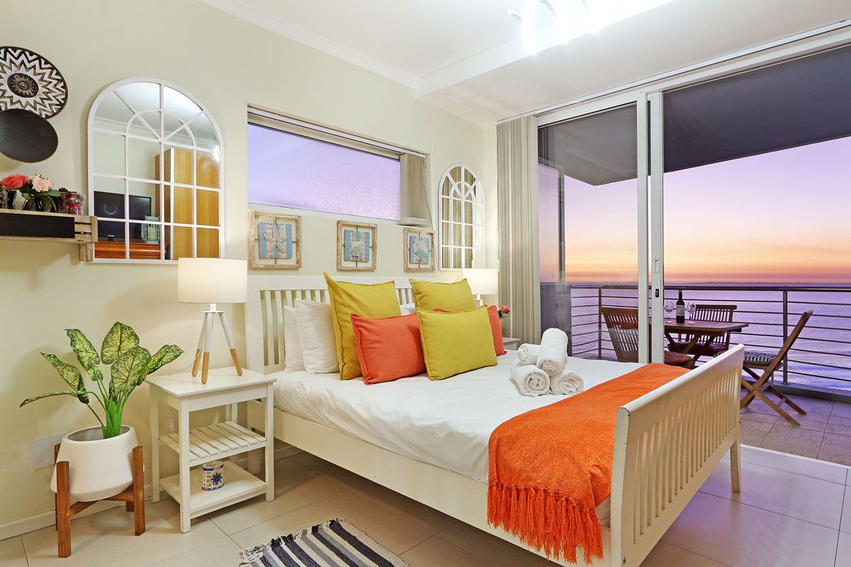A bedroom with a bed, bedside table, lamp, potted plant, and a view of the sunset through the balcony doors.