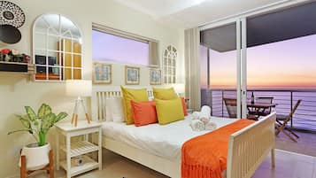 Ein Schlafzimmer mit Bett, Nachttisch, Lampe, Topfpflanze und Blick auf den Sonnenuntergang durch die Balkontüren.