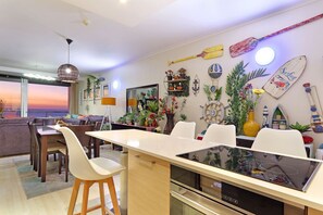A modern kitchen with a central island, dining area with white chairs, and a view of the sunset through large windows. - Horizon Bay 1201 (Cape Town)