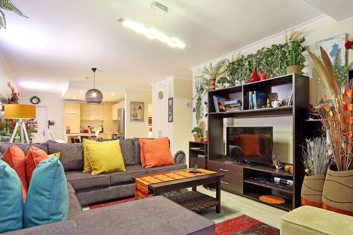 A modern living room with a grey sofa, colorful cushions, a coffee table, and a TV stand with a flat-screen TV. There is a bookshelf with decorative items and plants.