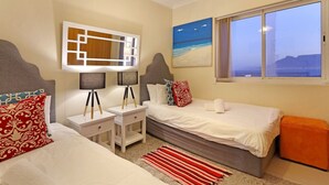 A hotel room with two beds, a nightstand with lamps, a painting of a beach, and a window with a view of the sea. - Horizon Bay 1201 (Cape Town)