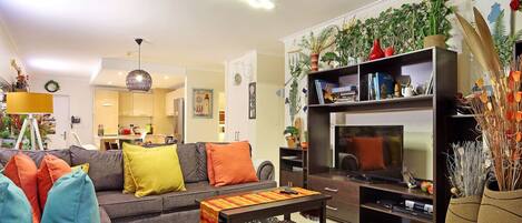 Uma sala de estar moderna com um sofá cinzento, almofadas coloridas, uma mesa de centro e um rack de TV com televisão de tela plana. Há uma estante com objetos decorativos e plantas.
