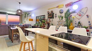 A modern kitchen with a central island, dining area with white chairs, and a view of the sunset through large windows. - Horizon Bay 1201 (Cape Town)
