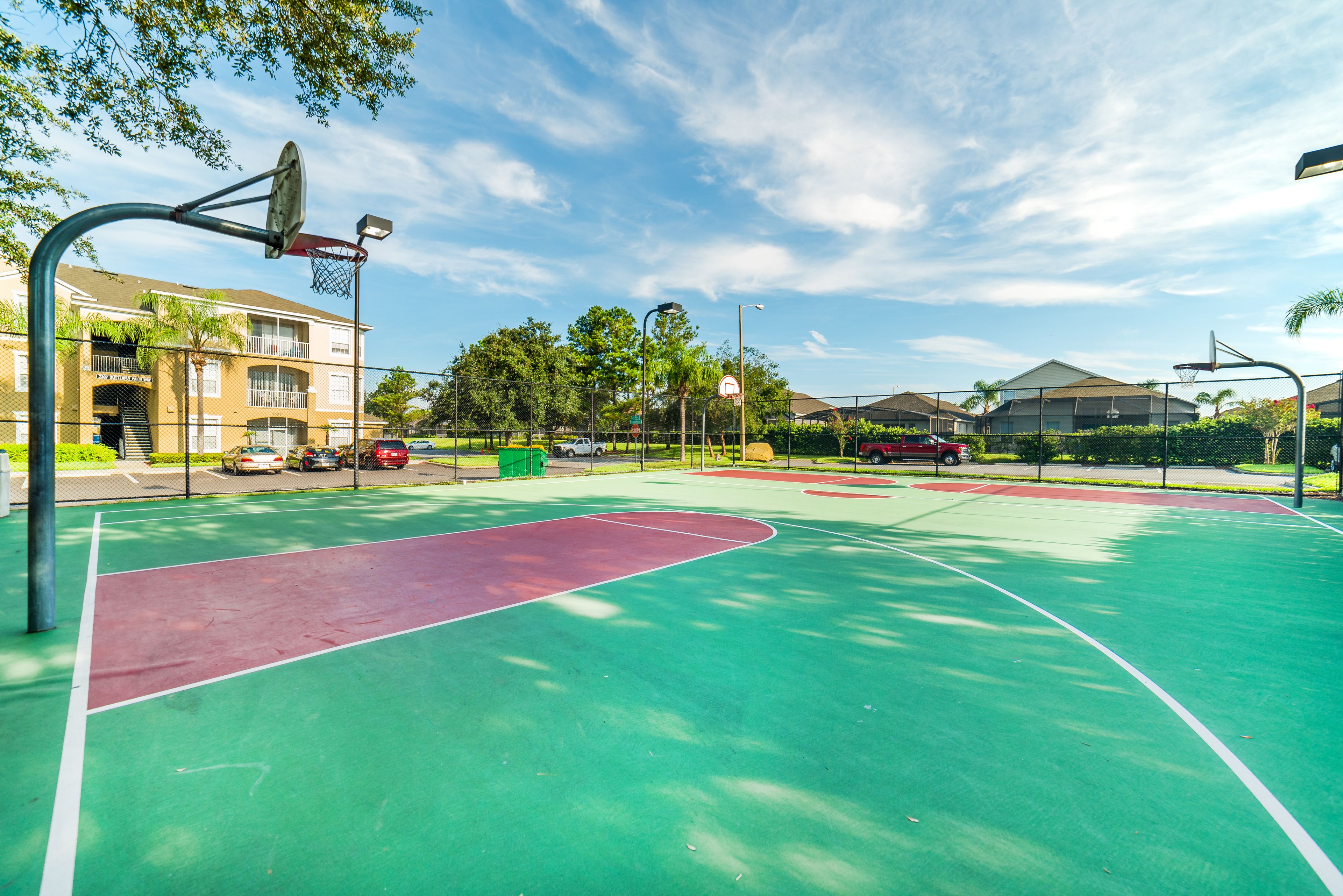 An outdoor basketball court with a green and red playing surface, surrounded by a fence, with residential buildings in the background.