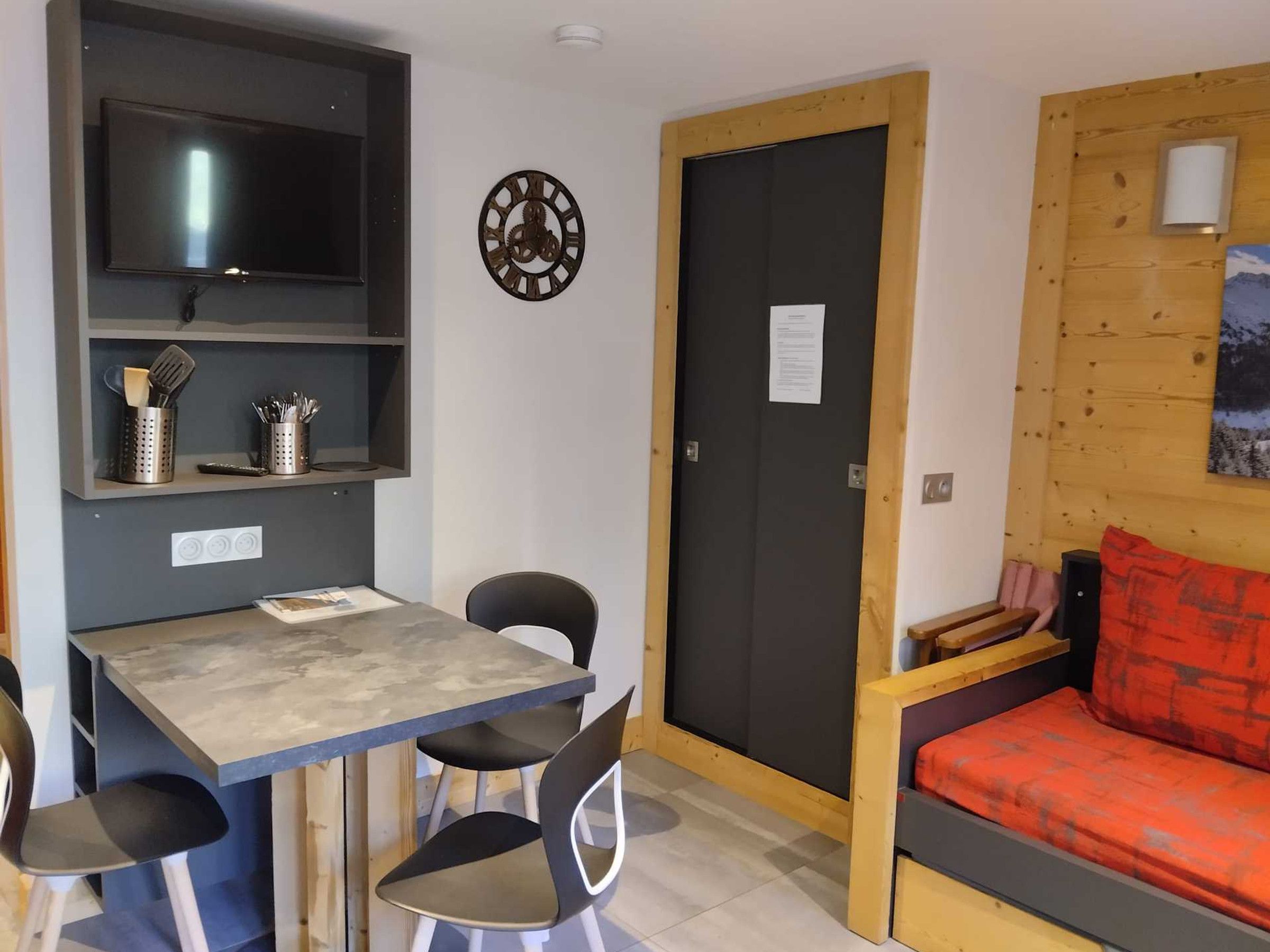 A compact room with a dining table, chairs, a television, a clock, a door, and a bed with a red cushion.