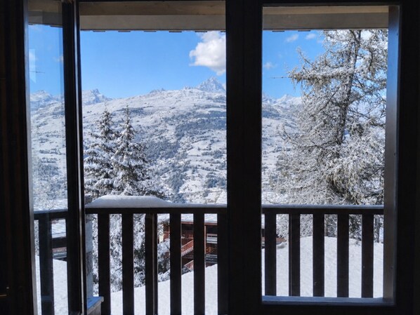 A view through a window with a wooden railing, overlooking a snowy mountain landscape with trees and a building in the foreground.