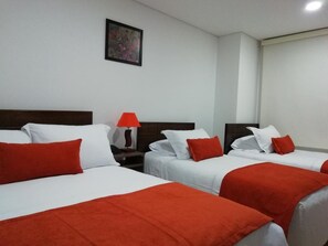 Quarto de hotel com duas camas, cada uma com roupa de cama branca e detalhes vermelhos, uma mesa de cabeceira com abajur vermelho e um quadro na parede.