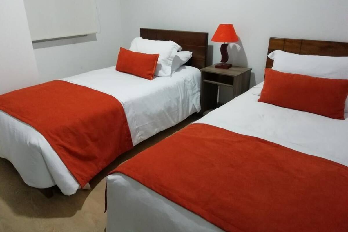 Two single beds with white and red bedding, a nightstand with a lamp, and a window with blinds.